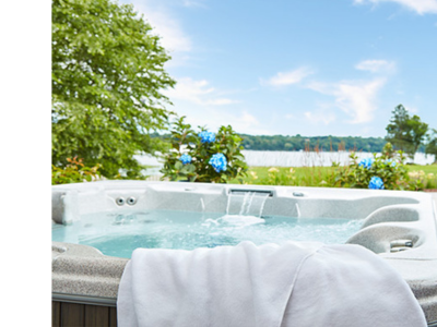 An outdoor hot tub is photographed with a lake and greenery in the background. The water is clear and there is a white towel hanging off the front edge of the spa.