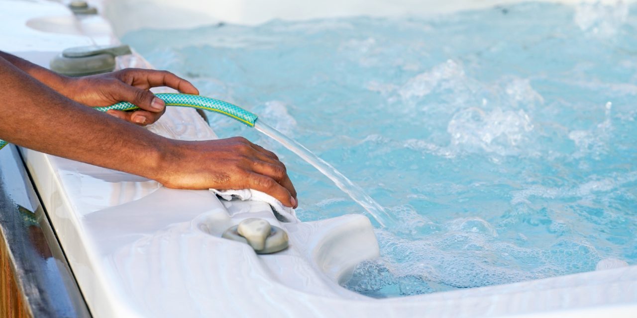 A man refills a swim spa with a green garden hose.