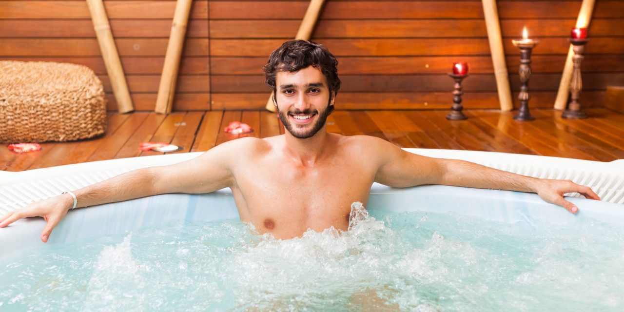 A man with longer hair and a beard relaxes in an indoor hot tub with his arms stretched out.