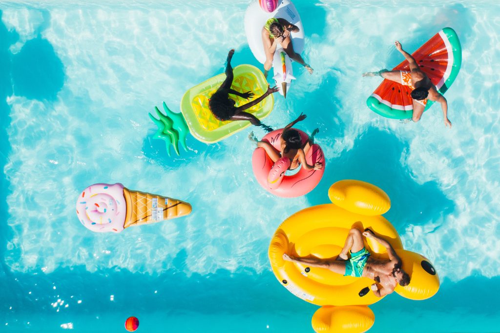 5 People sit on various pool floaties in an overhead shot of an outdoor pool