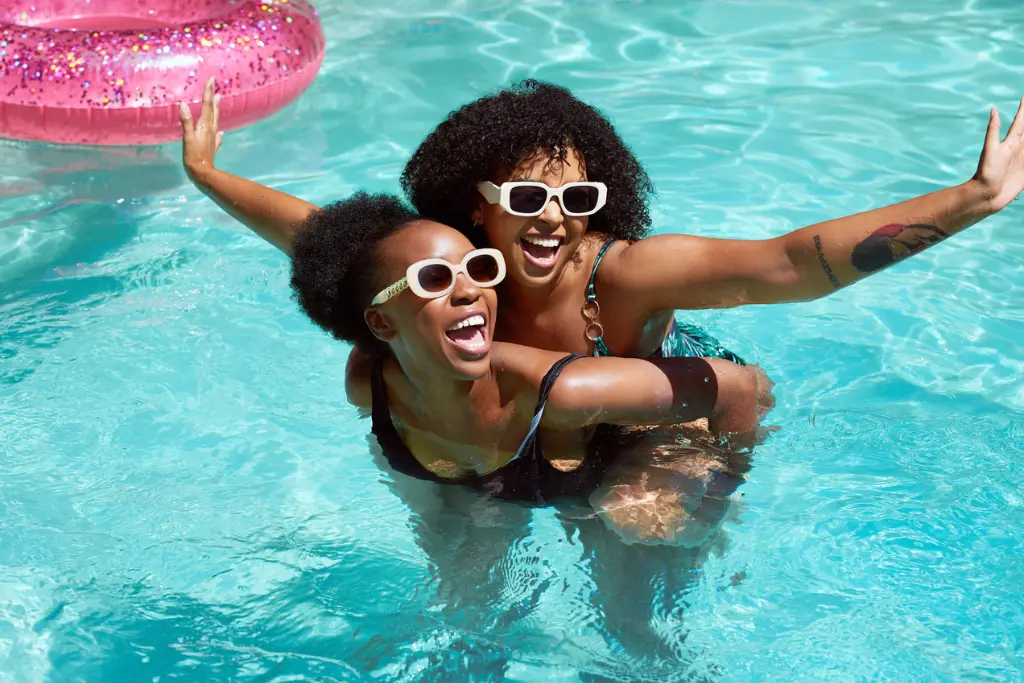 Two women are joyfully playing in a swimming pool on a sunny day. Both are wearing stylish white sunglasses and big smiles, with one giving the other a piggyback ride in the water. A sparkly pink pool float is visible in the background, and the clear turquoise water glistens in the sunlight, adding to the fun, carefree vibe of the scene.
