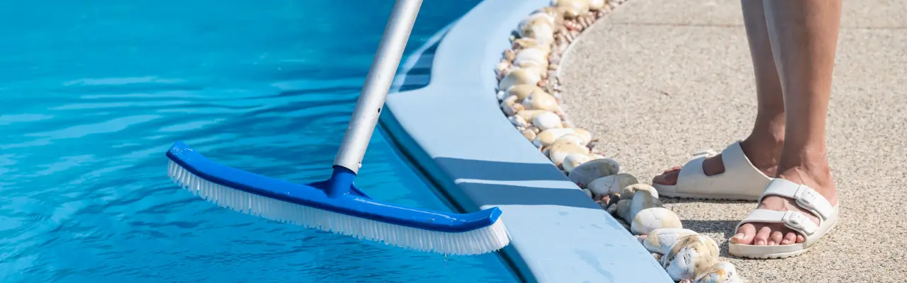 A person wearing white sandals stands at the edge of a swimming pool, holding a long-handled blue pool brush submerged in the water. White decorative stones line the pool’s edge.