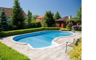 A backyard swimming pool surrounded by a neatly trimmed green hedge and stone tile patio, with a bench on one side. In the background are lush trees, flowering bushes, and houses with red-tiled roofs under a clear blue sky.