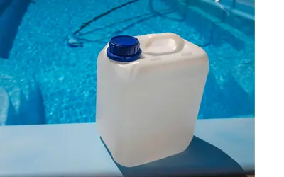 A white plastic container with a blue cap sits on the edge of a clear blue swimming pool, with pool cleaning tools visible in the water in the background.