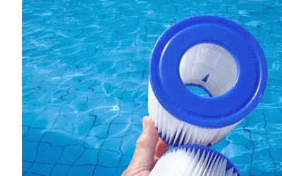 A hand is holding two cylindrical pool filters with blue rims and white pleated paper. The background shows a clear blue swimming pool with visible water ripples and a grid pattern on the pool floor.